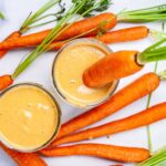 Two carrot and banana smoothies on a table and served with raw carrots.