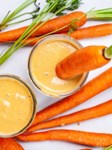 Two carrot and banana smoothies on a table and served with raw carrots.