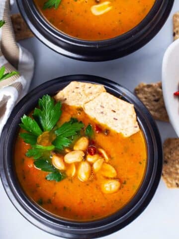 A closeup of a black bowl filled with simple carrot soup, topped with roasted peanuts, chili, fresh Parsley leaves, and rice crackers served next to a bushel of fresh Parsley and extra roasted peanuts.