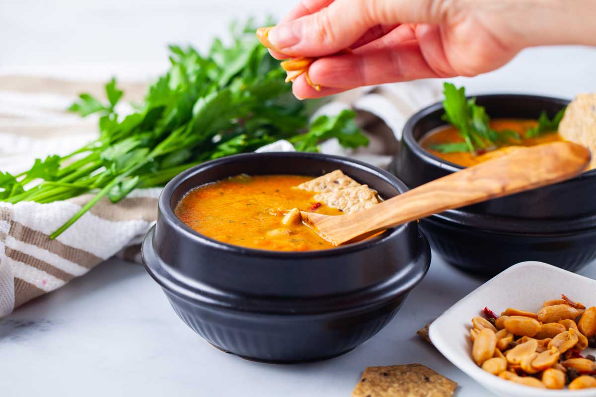 A closeup of a hand adding roasted peanuts as a topping to carrot soup served in a black bowl, next to a bushel of fresh parsley, and a small bowl of extra peanuts.