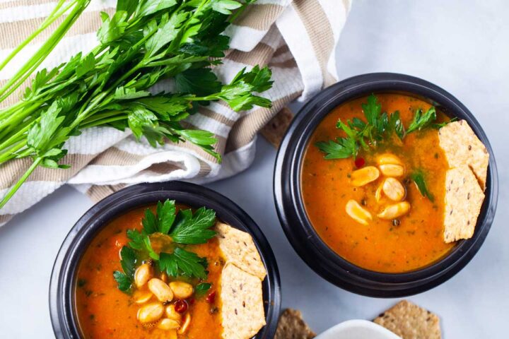 A closeup of two black bowls filled with pureed carrot soup, topped with roasted peanuts, chili, rice crackers, and fresh Parsley leaves, served next to a bushel of fresh Parsley.