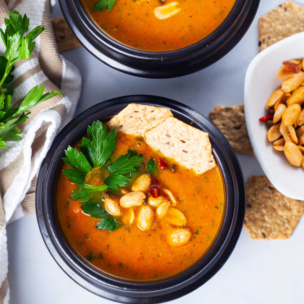 A closeup of a black bowl filled with simple carrot soup, topped with roasted peanuts, chili, fresh Parsley leaves, and rice crackers served next to a bushel of fresh Parsley and extra roasted peanuts.