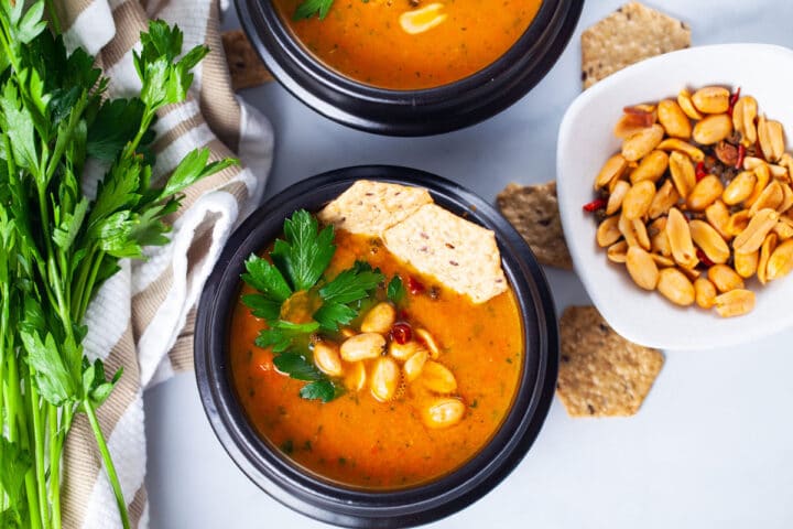 A closeup of a black bowl filled with simple carrot soup, topped with roasted peanuts, chili, fresh Parsley leaves, and rice crackers served next to a bushel of fresh Parsley and extra roasted peanuts.