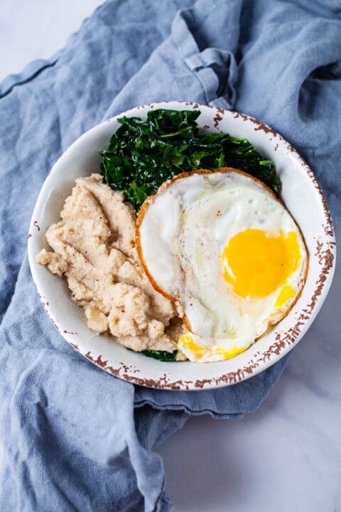 A bowl filled with mashed cauliflower, sauteed kale, and egg sunny side up.