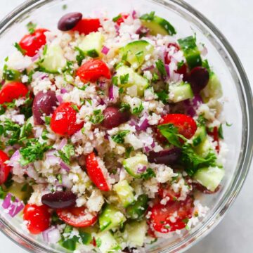 Shredded cauliflower with cucumbers, mini tomatoes, onions, and olives in a bowl.