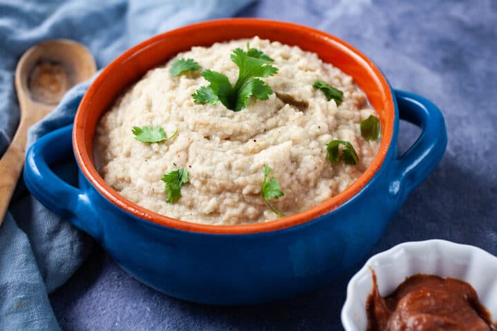 A terra cotta bowl filled with creamy mashed cauliflower and garnished with Parsley, next to a small bowl with apple butter.