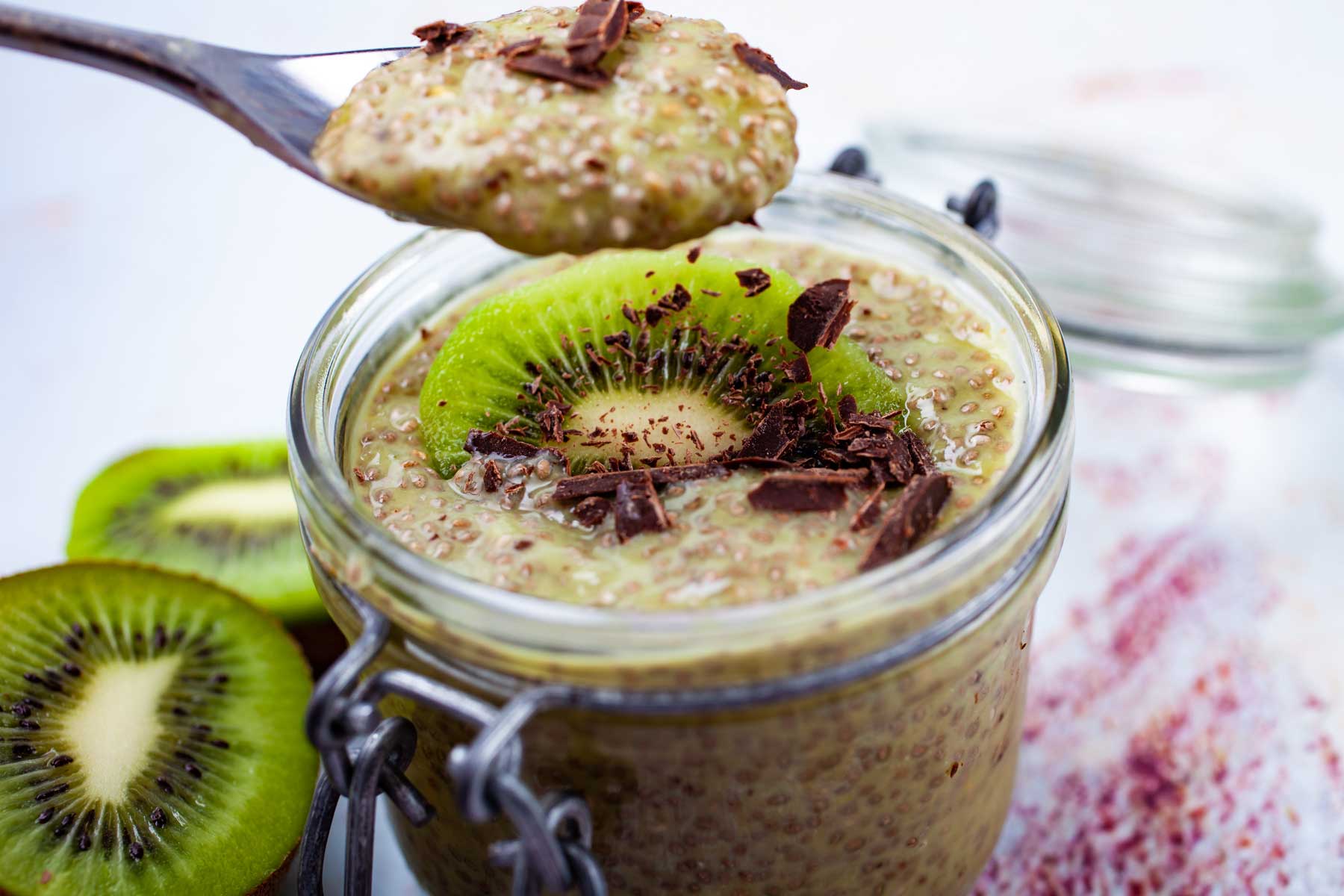 A hand holding a full spoon of kiwi chia pudding topped with shaved dark chocolate.