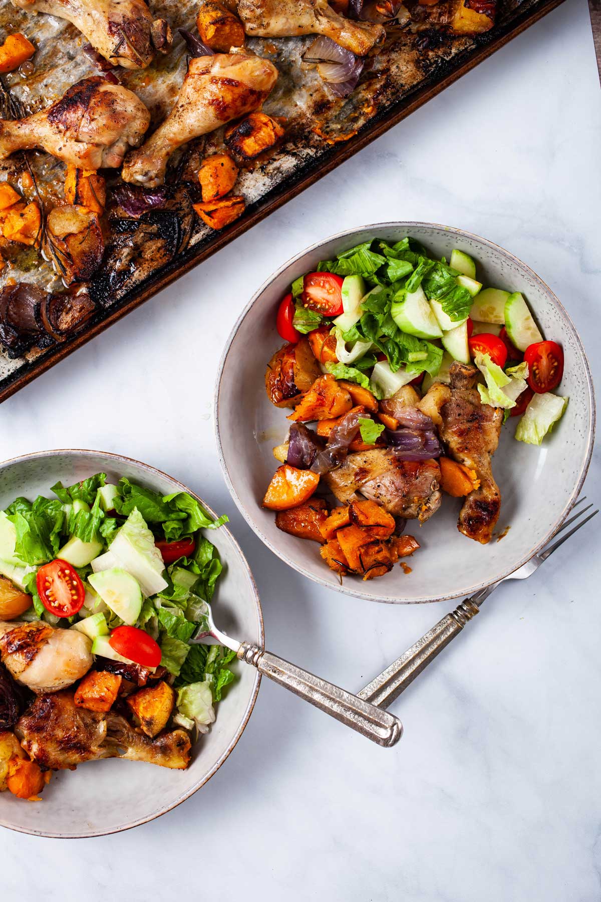 Two plates filled with roasted drumsticks, sweet potatoes, apples and a green salad next to a chicken dinner sheet pan.