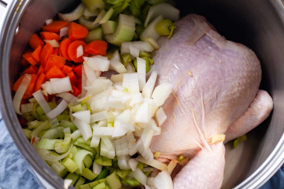 A large pot filled with a raw large chicken, and fresh chopped vegetables from carrots, onions, leeks, and celery.