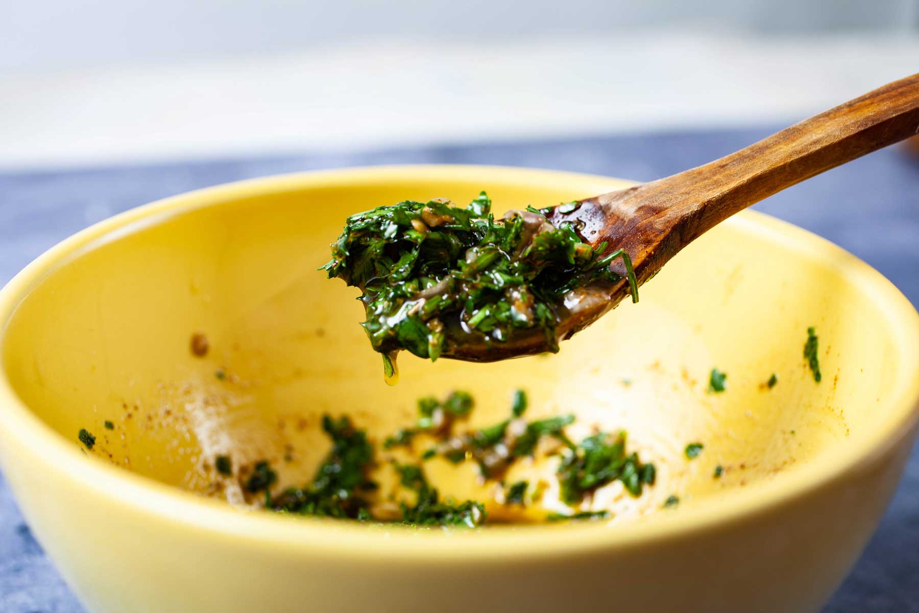 A wooden spoon lifted over a bowl with chimichurri sauce.