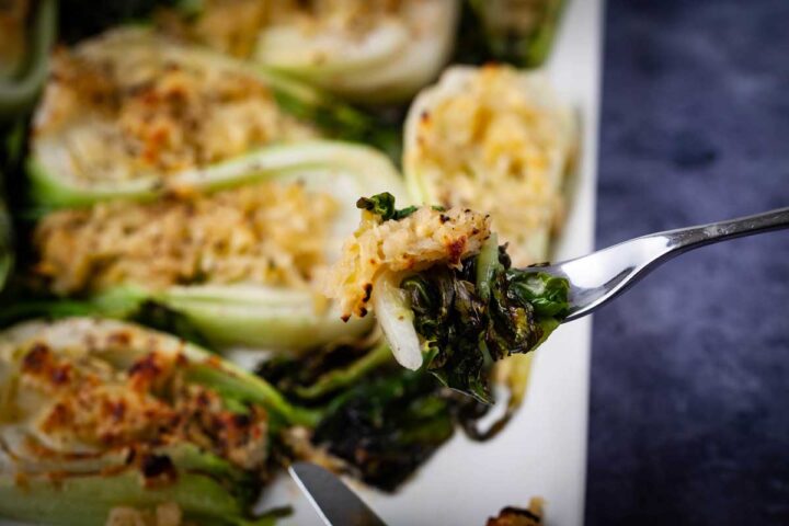 A close up of a fork holding up a bite of cheesy roasted baby bok choy.