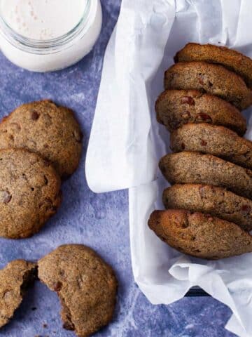 A basket filled with buttery buckwheat cookies, next to a cup of milk and some cookies spread out on the table.