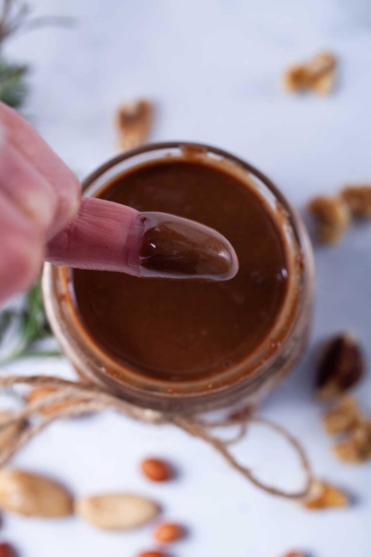 A finger topped with glossy chocolate peanut butter with the nut butter jar in the background.