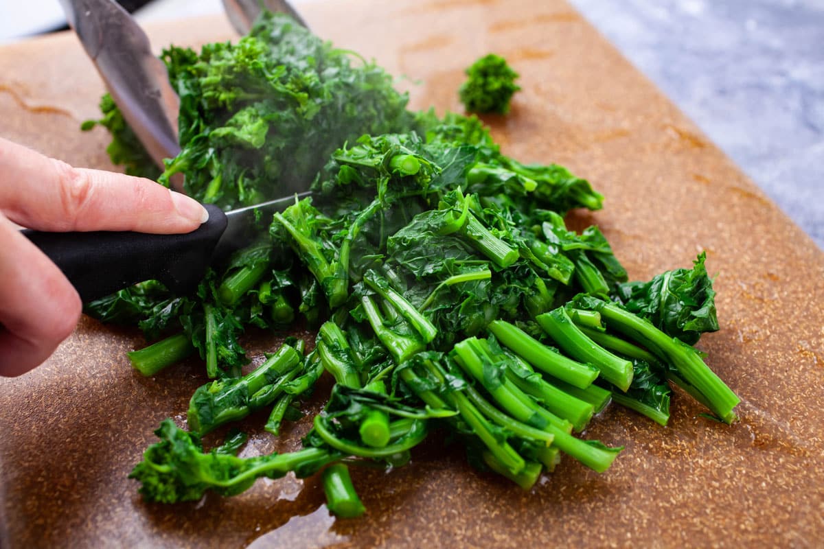 Someone cutting cooked broccoli rabe on a cutting board.