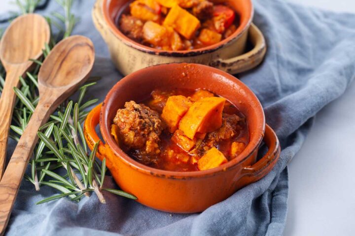 Two terracotta bowls filled with a meat sweet potato and stew, served next to two wooden spoons and fresh Rosemary leaves.