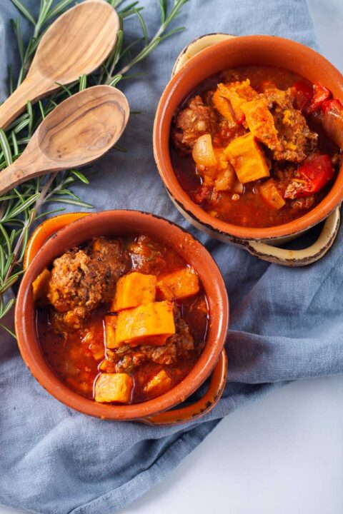 Two terracotta bowls filled with a meat sweet potato and stew, served next to two wooden spoons and fresh Rosemary leaves.