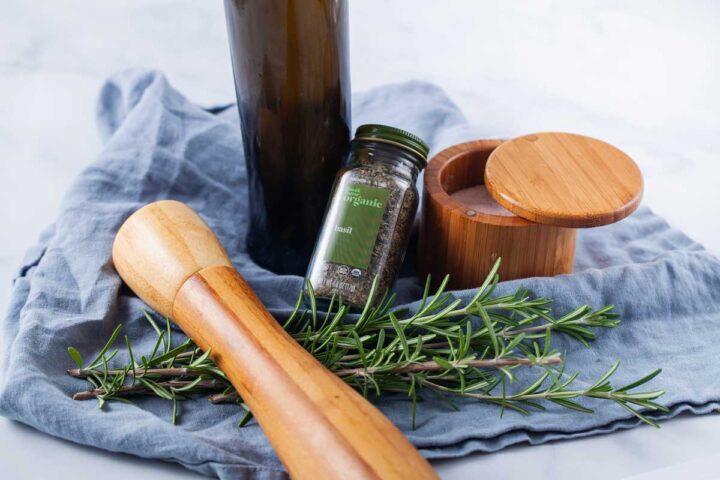 A wooden pot filled with salt, dried basil seasoning, a wooden pepper mill, an olive oil bottle, and fresh Rosemary leaves showcased on a blue kitchen towel.