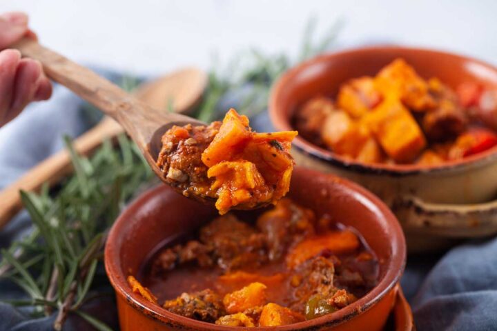 A close-up of a wooden spoon taking a serving of meat and sweet potato stew out of a single serving bowl.