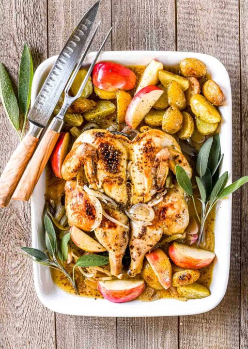 an overhead shot of a spatchcocked chicken in a white casserole dish filled with apples and potatoes and garnished with large sprigs of fresh sage.