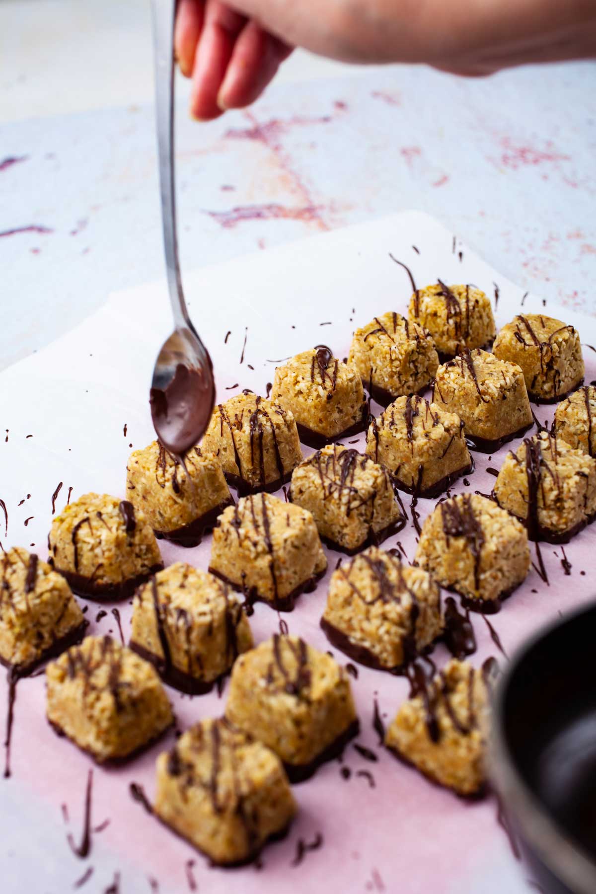 A hand holding a chocolate covered spoon to create zig-zagging chocolate streaks over coconut bites.