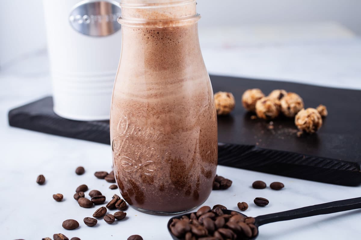 A coffee date smoothie in a small glass jar surrounded by coffee beans and oat snacks.