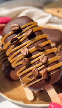 A hand holding a coffee ice cream on a stick topped with caramel and coffee beans.