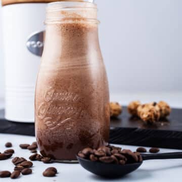 A chocolate coffee smoothie in a small glass jar surrounded with coffee beans and oat snacks.