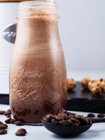 A chocolate coffee smoothie in a small glass jar surrounded with coffee beans and oat snacks.