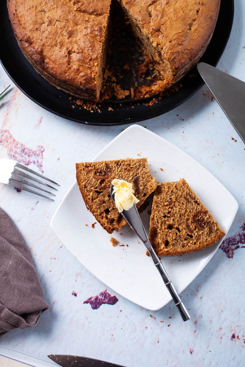 A slice of fig cake cut open and resting on a plate next to the cake and topped with butter.