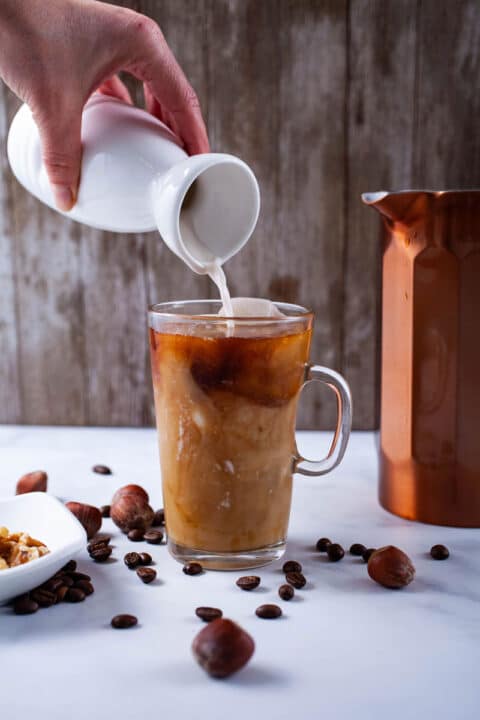 A hand pouring milk with a milk can into an iced glass coffee cup.