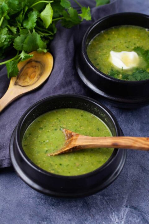 Two black bowls filled with green pea soup, one topped with fresh Parsley leaves and sour cream, the other containing a wooden spoon resting in the bowl.