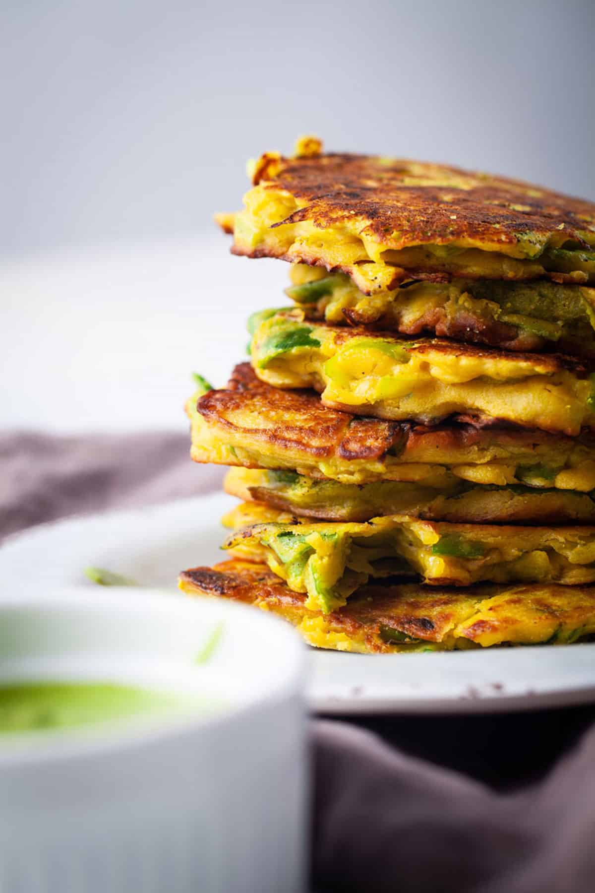 A stack of crispy leek veggie cakes served on a plate with a pesto sauce on the side.