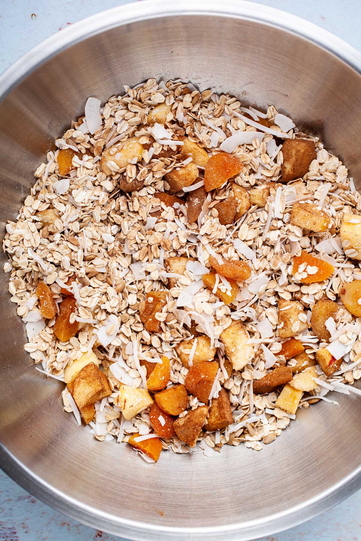 Oats, seeds, apples, dried fruits, coconut flakes, and spices combined in a large mixing bowl.