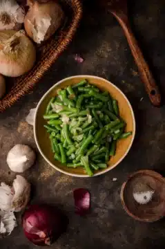 Green beans and onions in a bowl, surrounded by spices, garlic, and onions.