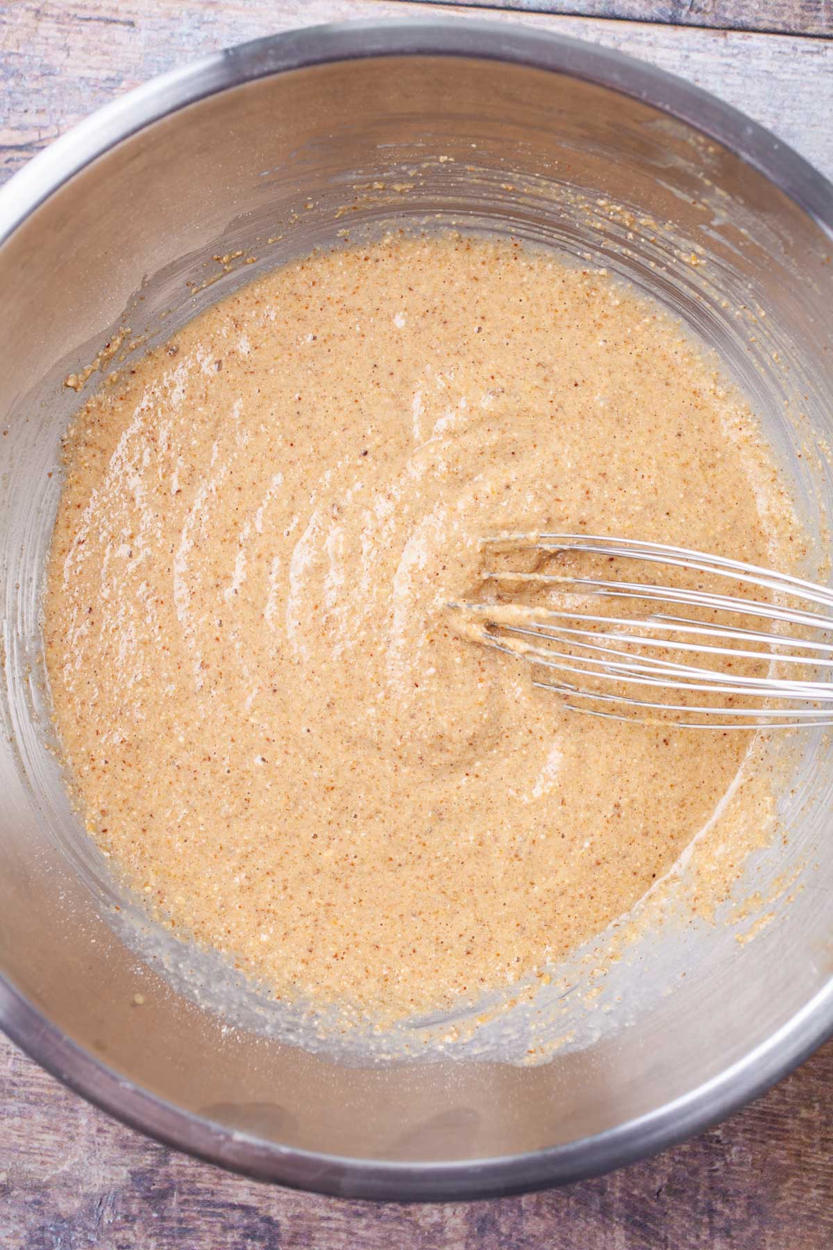 A whisk is resting in pancake batter. in a stainless steel bowl.