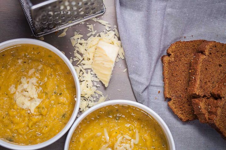 Two bowls filled with yellow squash soup, topped with Parmesan cheese and served with extra cheese and bread on the side.