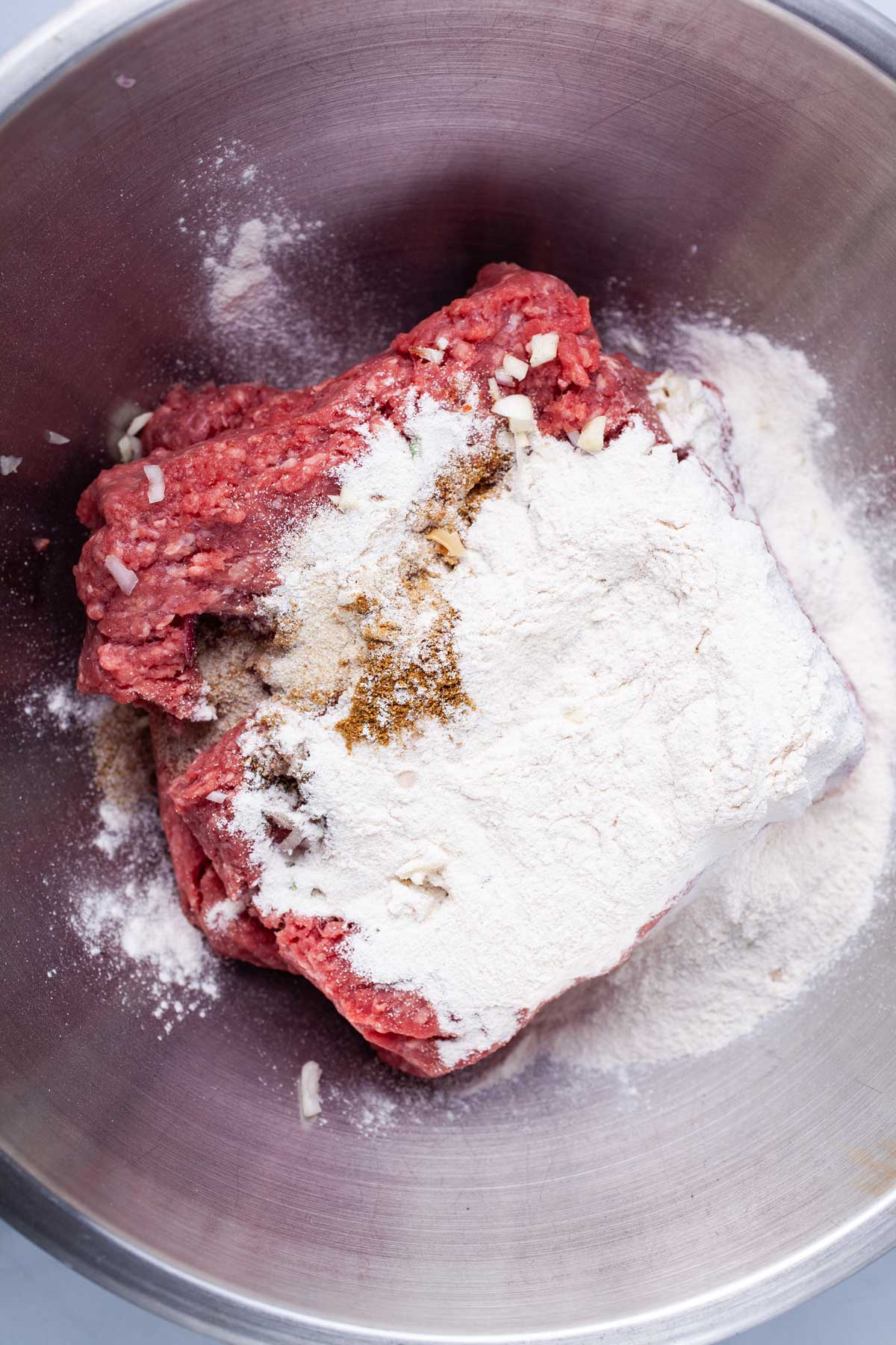 Flour and spices on top of raw ground meat in a stainless steel bowl.