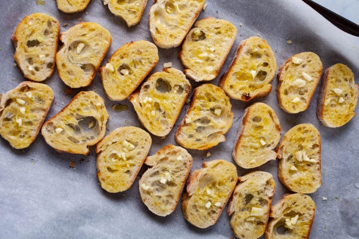 Slices of baguette bread displayed on an oven pan and topped with olive oil and freshly chopped garlic.