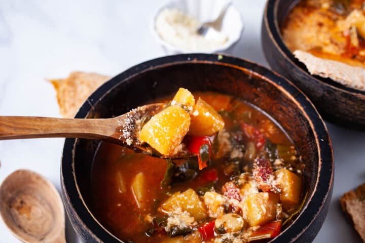 A wooden spoon with hearty veggies held above a soup bowl.