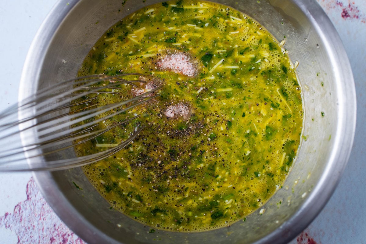 A whisk resting in a bowl with mixed eggs, fresh parsley, cheese, and seasoning.