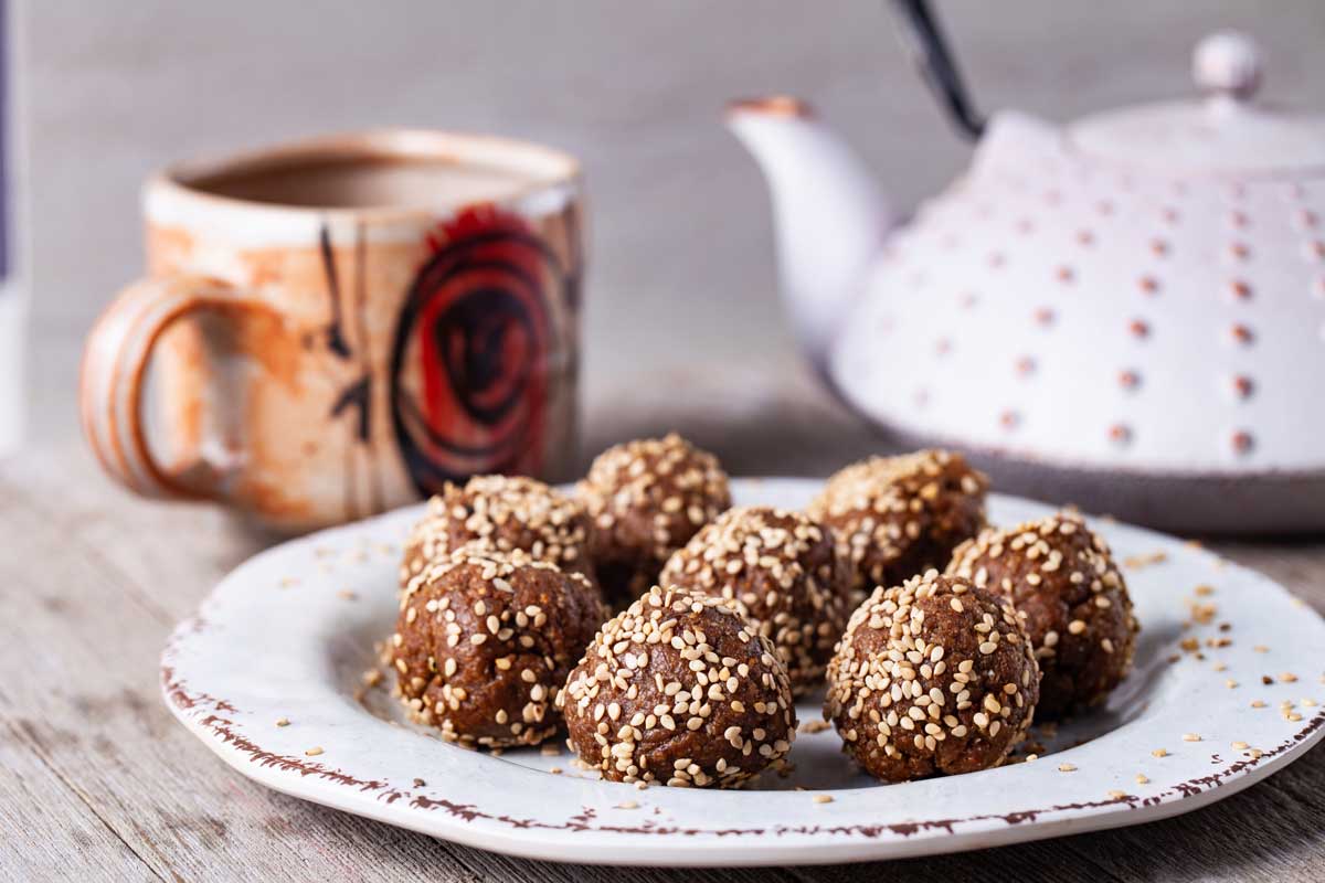 Energy balls sprinkled with seeds served on a small plate next to a cup and Asian teapot.