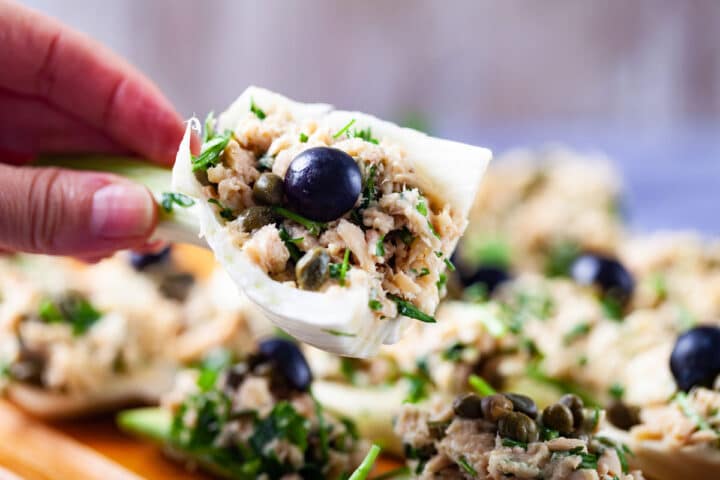 A hand holding a part of a raw fennel bulb filled with creamy tuna salad and garnished with a dark grape.