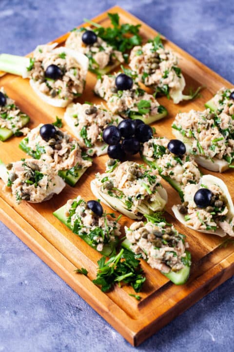 Fresh cucumber and fennel slices topped with tuna salad and garnished with dark grapes neatly presented on a wooden board.