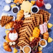 Fresh fruits, cheeses, charcuterie, meats, chocolate, croissants, spreads, coffee, juices, nuts, and homemade waffles all beautifully displayed on a waffle board and table.