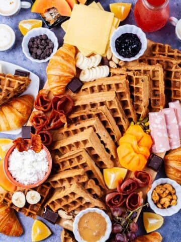 Fresh fruits, cheeses, charcuterie, meats, chocolate, croissants, spreads, coffee, juices, nuts, and homemade waffles all beautifully displayed on a waffle board and table.