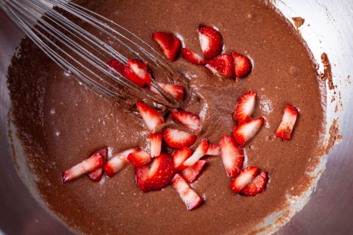 A whisk resting in flourless cake batter with fresh chopped strawberries in a bowl.