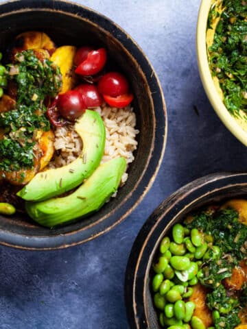 Bowls filled with cooked rice, small tomatoes, avocado slices, fried edamame beans, fried plantains, and topped with chimichurri sauce.