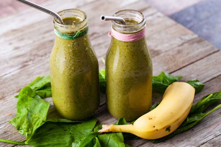 Two milk bottles filled with green juice, surrounded by half a banana and fresh spinach.