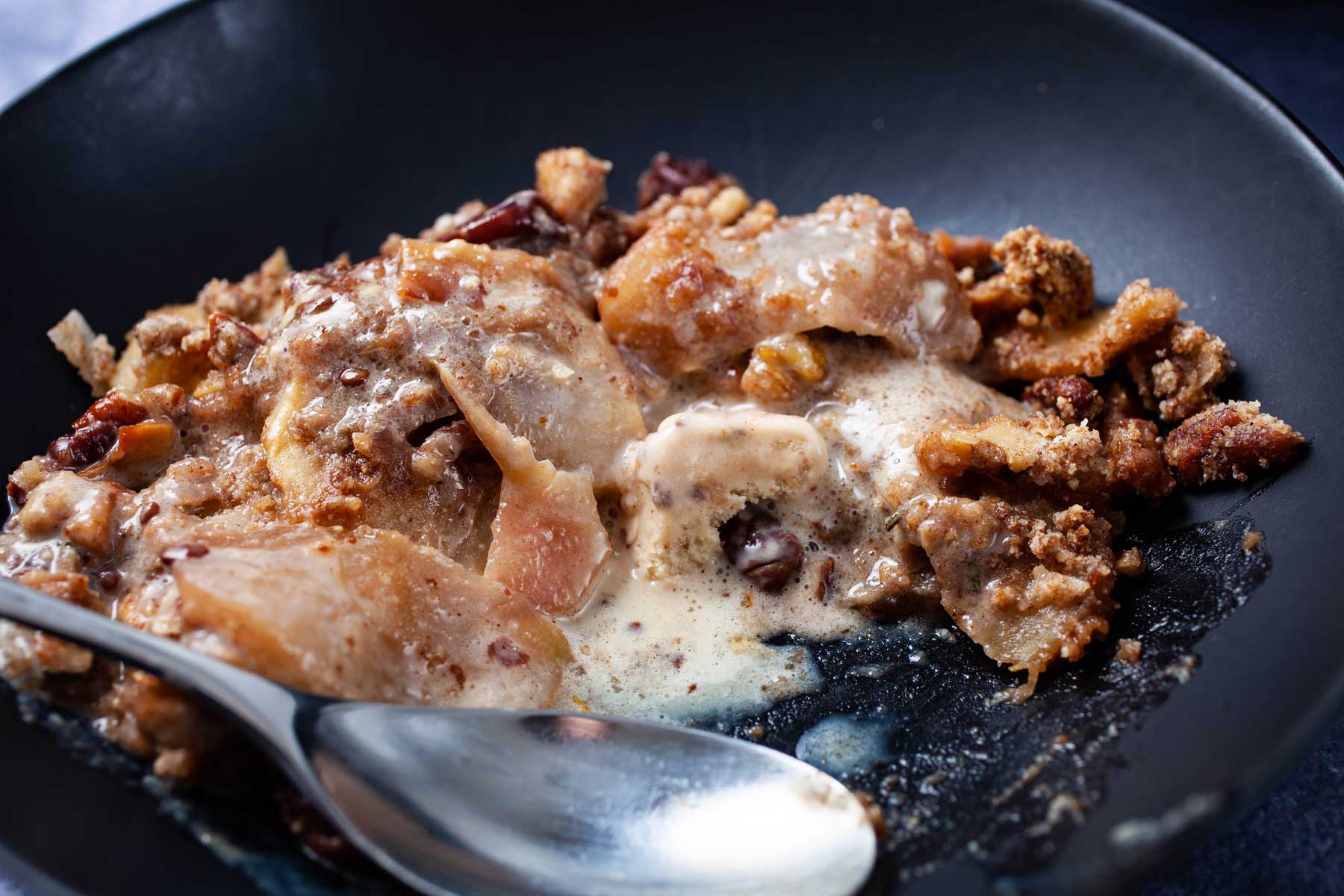 A large spoon resting on a plate with ice cream melted on baked apple crumble.