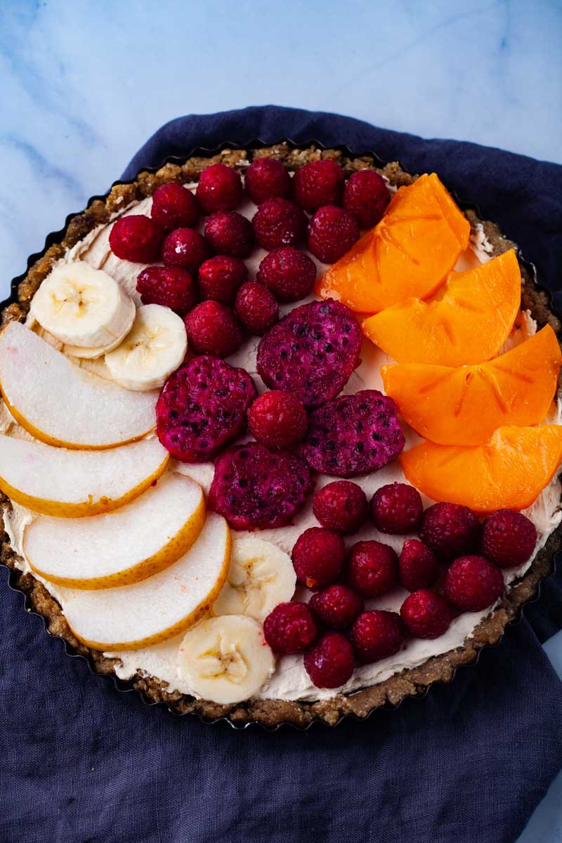 A round tart with a no bake crust, cheese filling, and topped with fruits like Asian peers, bananas, persimmon, raspberries, and red dragon fruit.